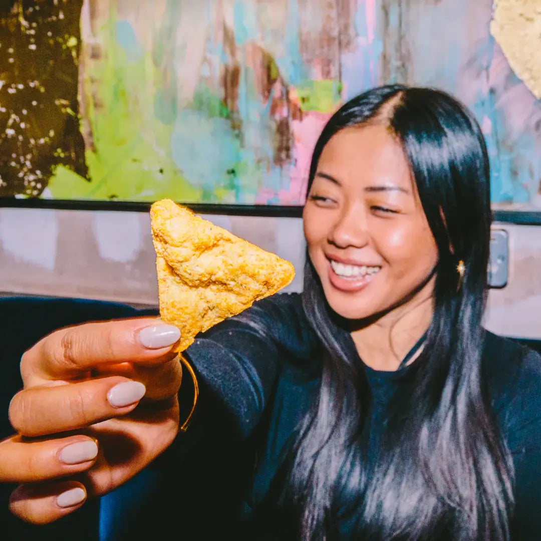 Smiling woman holding up a single triangular high-protein chip from The Curators, ready to snack